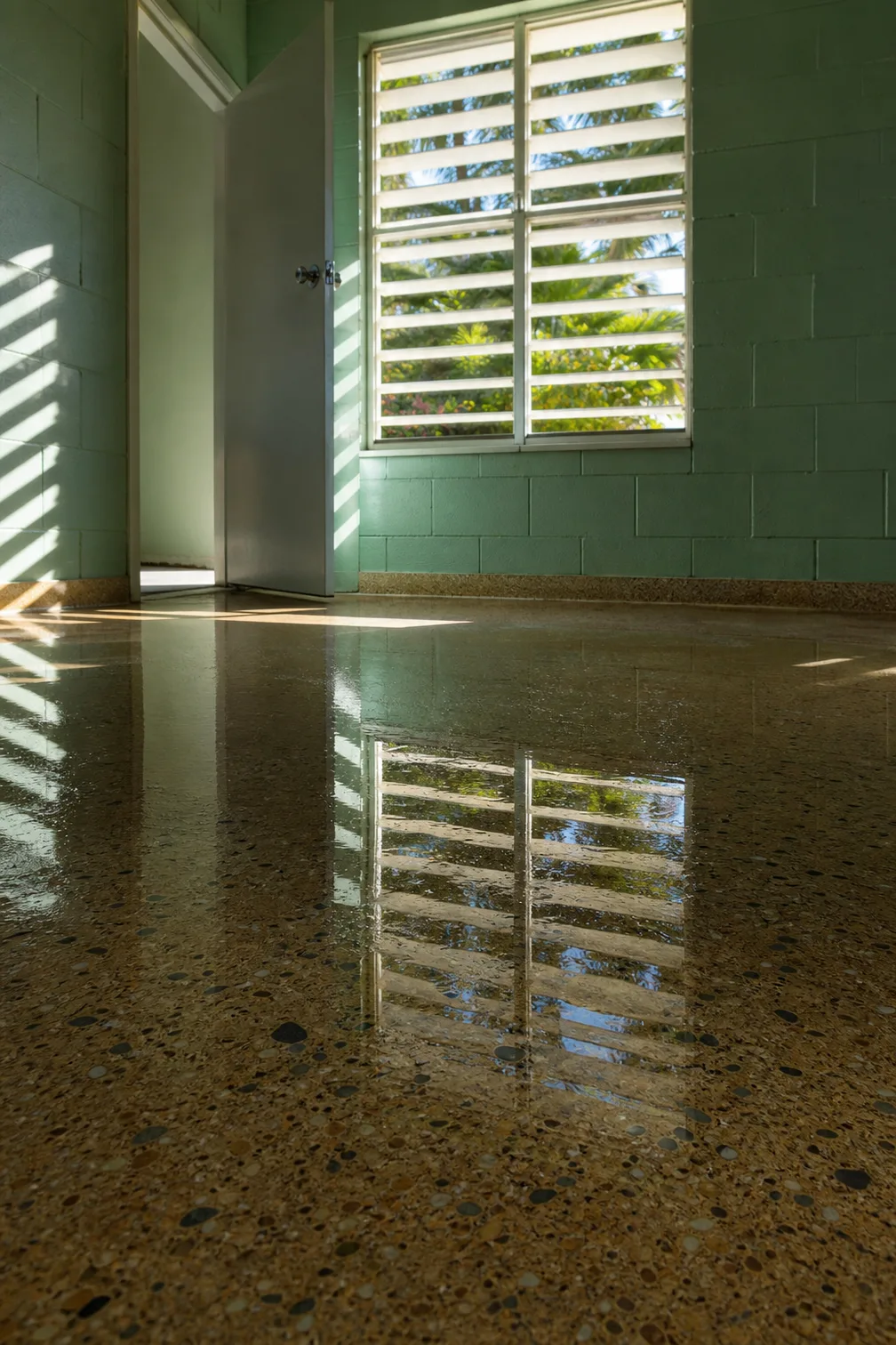 A wide shallow sheet of standing water across a polished terrazzo floor in an empty painted block room, holding the reflection of a louvred window