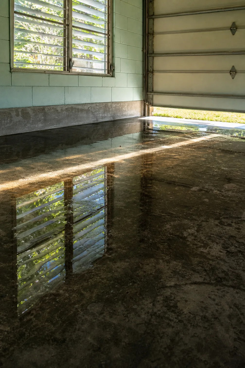 The inside of a garage with the up-and-over door raised a foot, daylight lying in a low band across a wet concrete slab