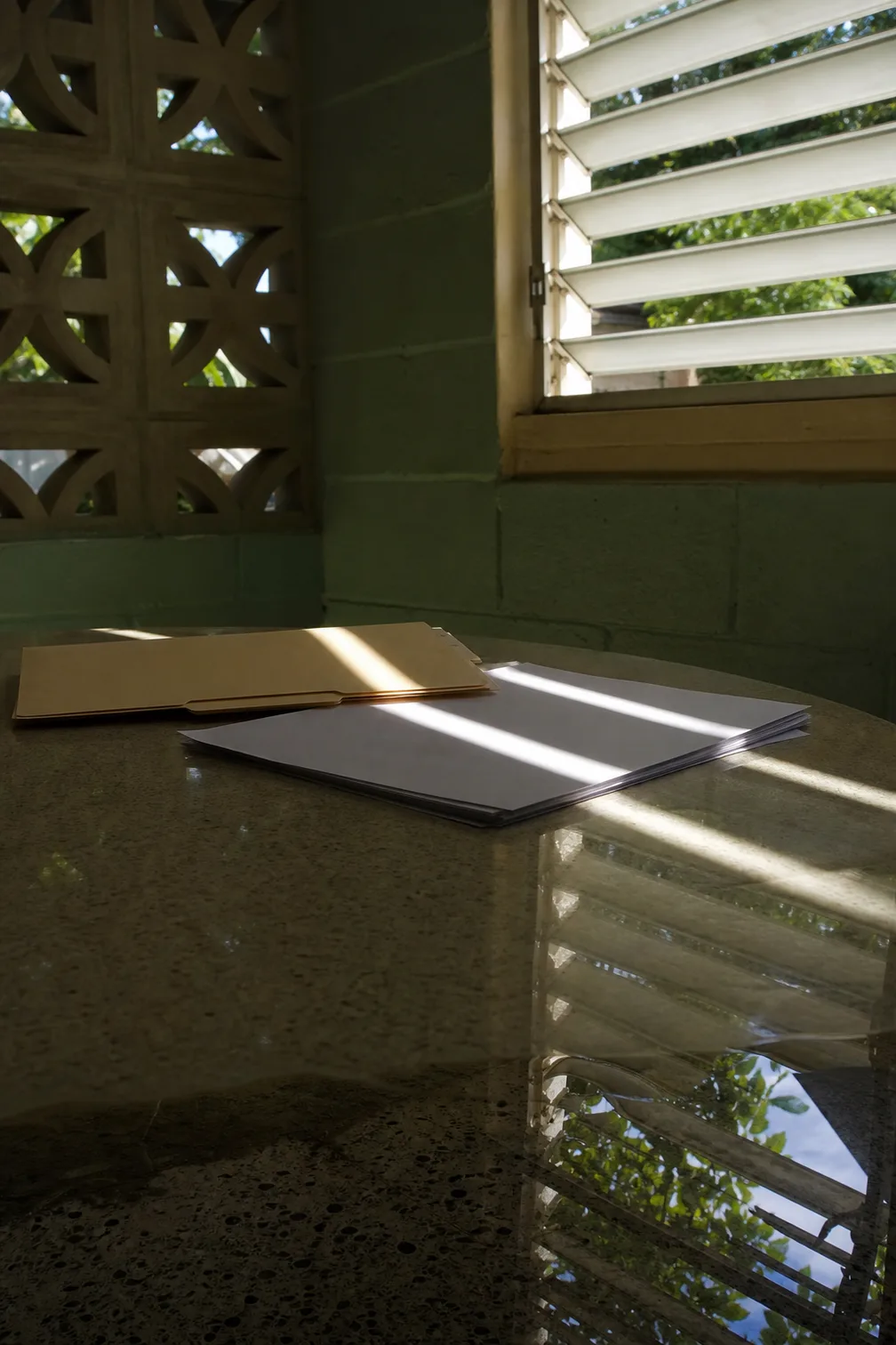 A manila folder and a stack of blank paper on a round terrazzo table, lit in bands by a jalousie window that is reflected in the polished top
