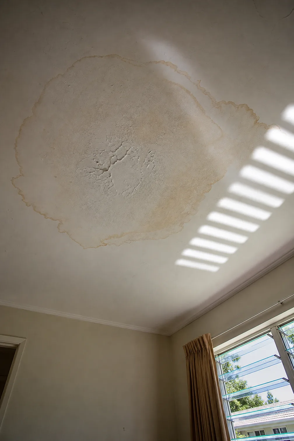 Brown water ring spreading across a flat white ceiling beside a hard band of sunlight